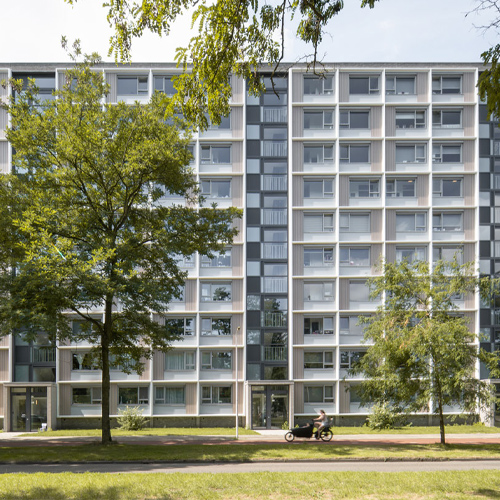 Exterior view of a renovated mid-rise residential apartment building with a grid-like façade of white-framed windows and balconies, flanked by green trees, with a cyclist passing on the street in front. Architecture by DOOR-architecten.