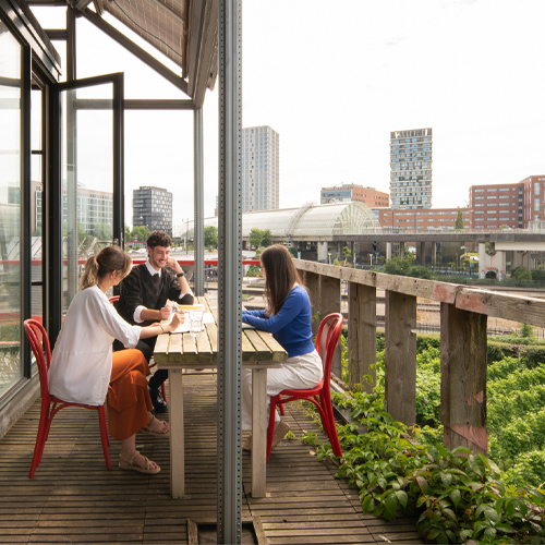 Drie collega's overleggen aan een houten tafel op een begroeide buitenterras met rode stoelen, met op de achtergrond een stedelijke skyline en een spoorwegviaduct op een bewolkte dag. Werksfeerbeeld bij DOOR-architecten.