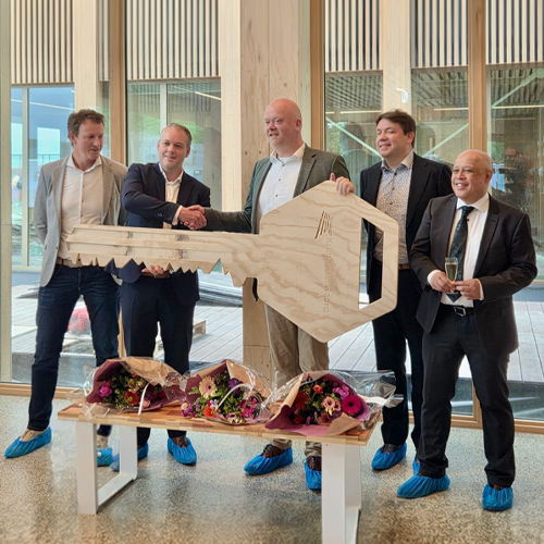 Vijf mannen poseren bij een officiële opleveringsceremonie in een modern gebouw, waarbij twee van hen een grote houten sleutel vasthouden en een handdruk geven, met bloemenboeketten op de voorgrond. Oplevering door DOOR-architecten.